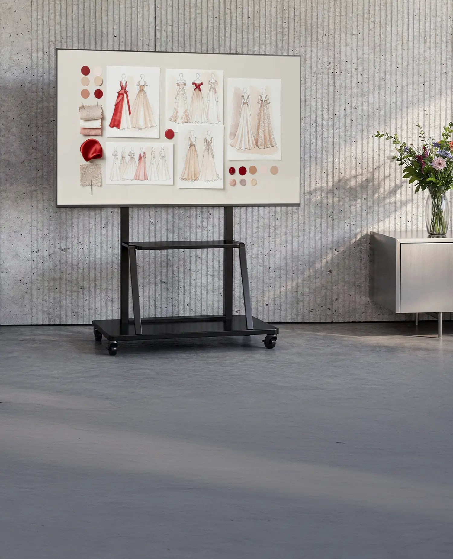 The image shows a modern room with a concrete wall and minimalist furnishings. At the center stands a mobile presentation display on a celexon Economy height adjustable display trolley Adjust-55110MP, showing a mood board with fashion and design concepts, fabric samples, and color palettes. Next to it is a sideboard with a vase full of flowers, creating a creative studio or design workspace atmosphere.