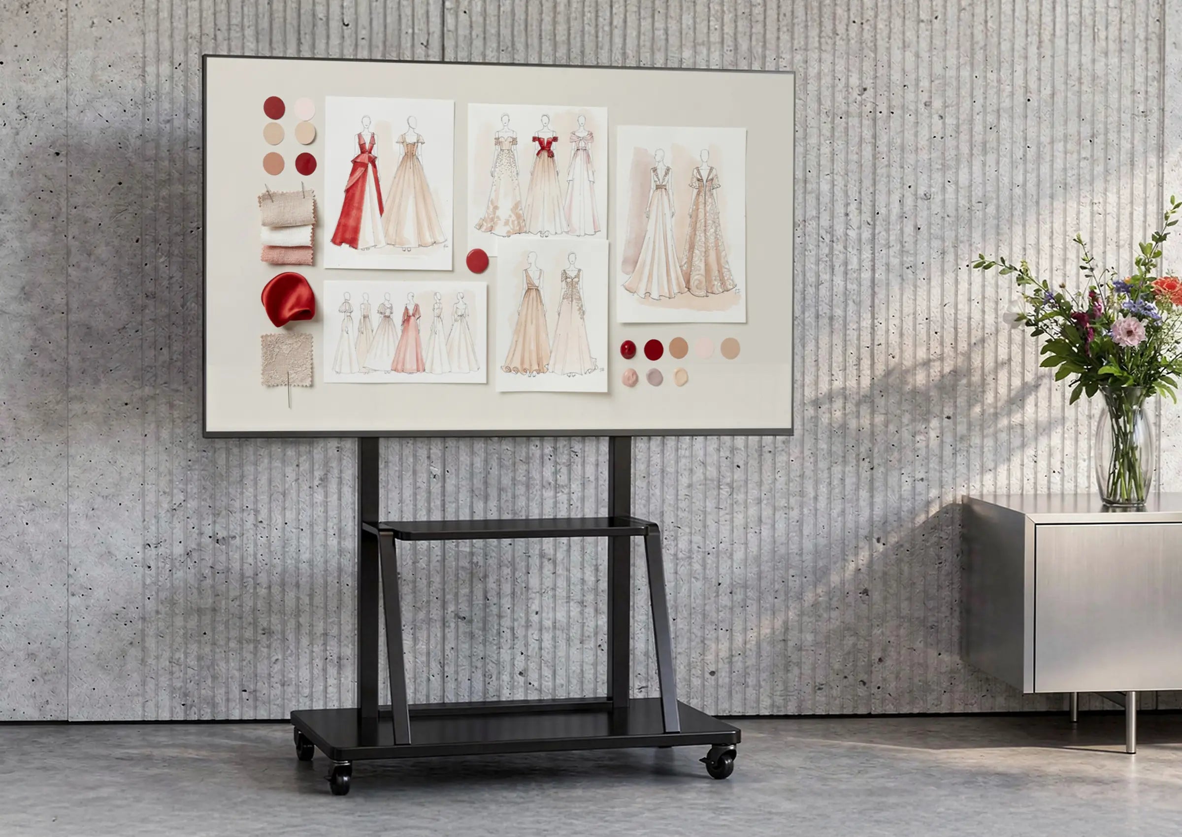 The image shows a modern room with a concrete wall and minimalist furnishings. At the center stands a mobile presentation display on a celexon Economy height adjustable display trolley Adjust-55110MP, showing a mood board with fashion and design concepts, fabric samples, and color palettes. Next to it is a sideboard with a vase full of flowers, creating a creative studio or design workspace atmosphere.