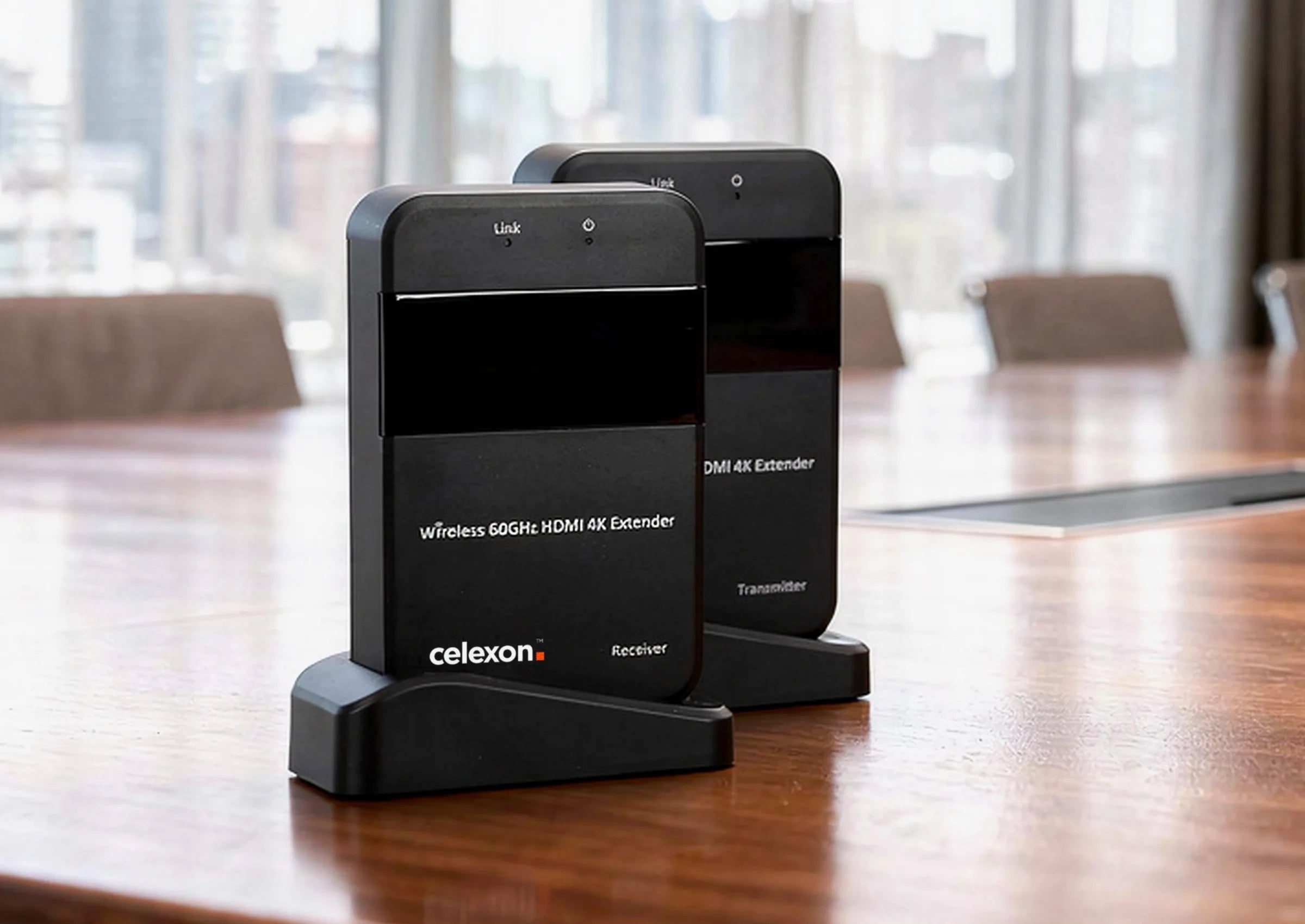 In the foreground, a celexon Expert Wireless HDMI Set WHD30M – 4K is placed on a large wooden table in a modern conference room, shown clearly and sharply. In the softly blurred background, two business professionals are conversing in front of floor-to-ceiling windows with a city view, while a presentation display is visible on the wall.