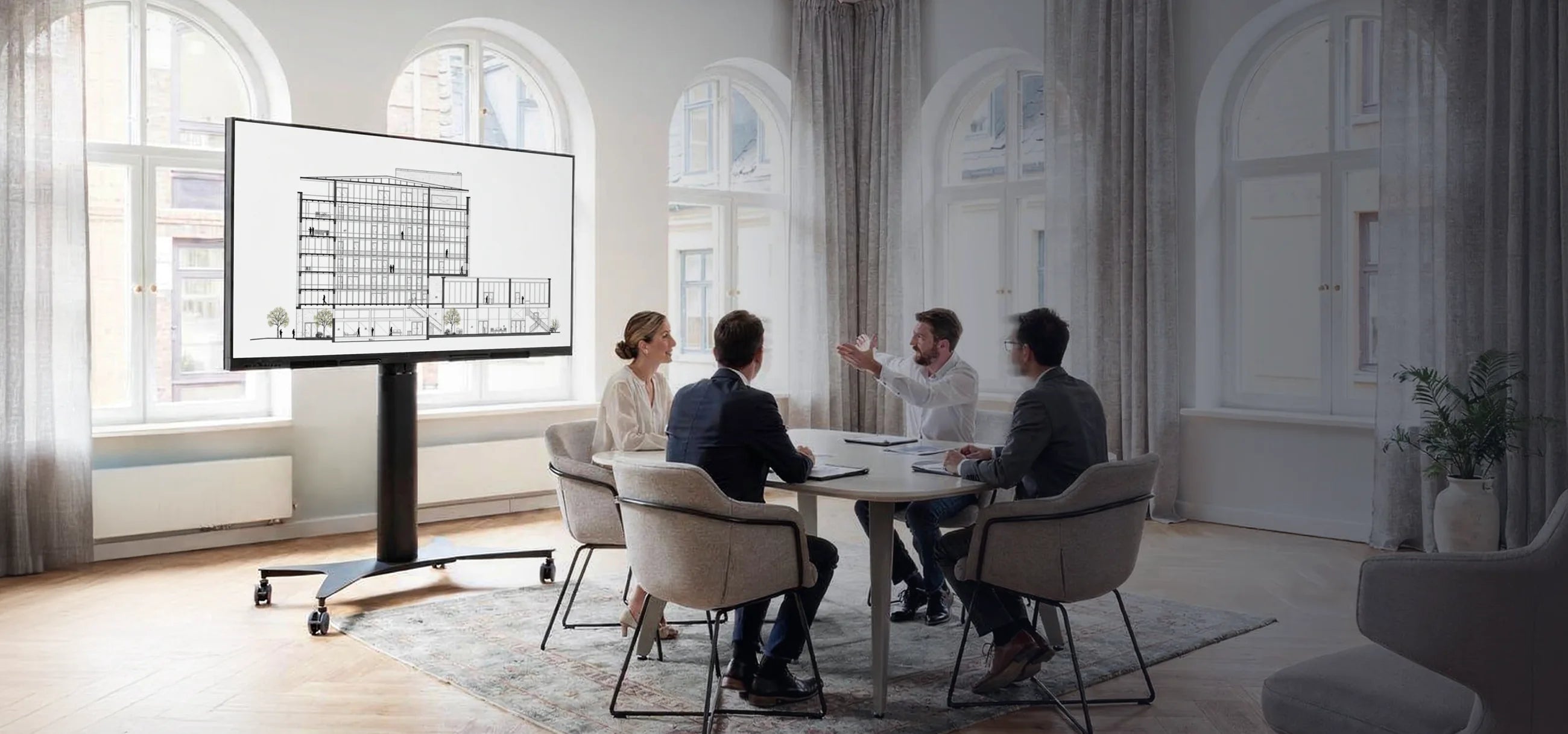 Four people are seated at a table in a bright, modern room, holding a meeting. On the left stands a celexon Expert electrically height-adjustable display trolley Adjust 42100M, showing an architectural drawing.