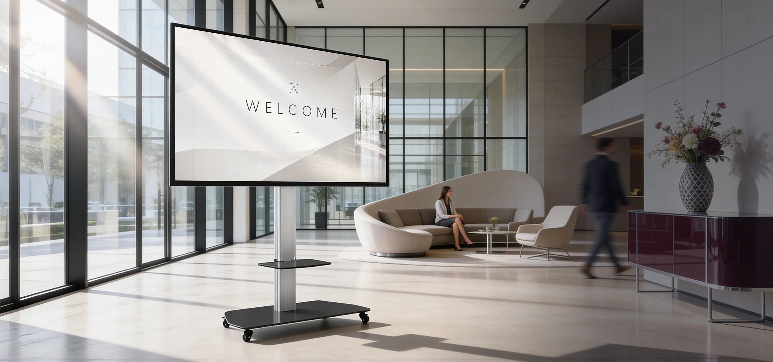 A bright, modern lobby with large glass fronts and elegant seating furniture; in the foreground stands a mobile digital display on a celexon Professional height-adjustable display trolley Adjust-3270MP with the word “Welcome” on it. In the background, people are sitting and walking through the open reception area with stylish furnishings and floral decorations.