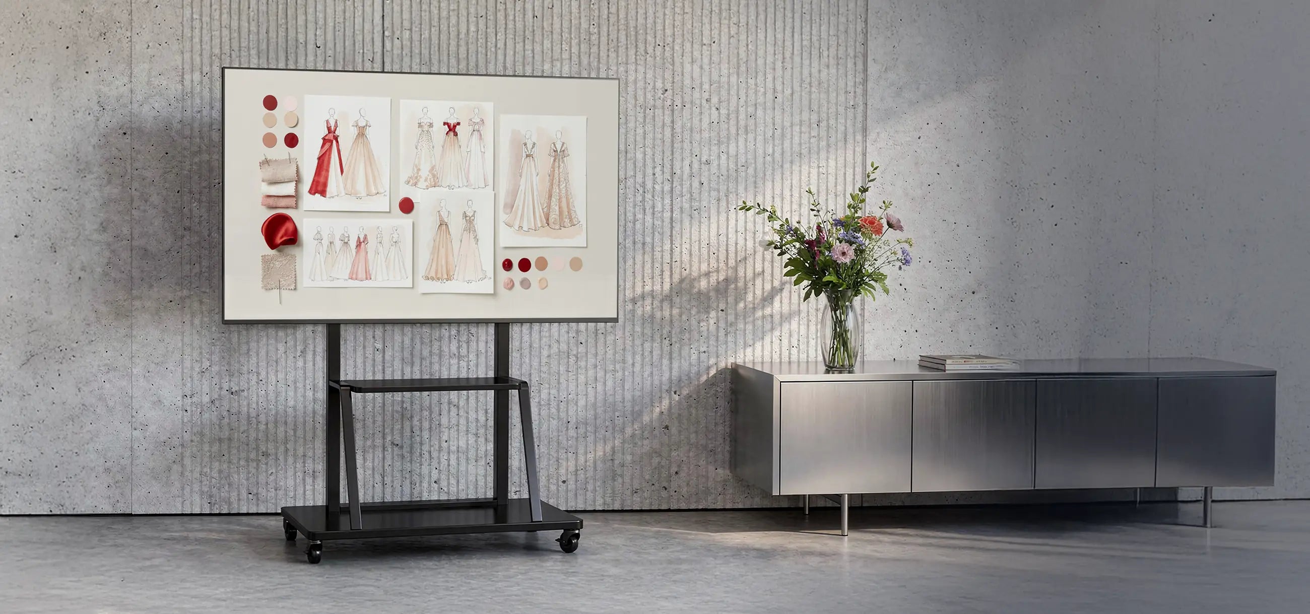 The image shows a modern room with a concrete wall and minimalist furnishings. At the center stands a mobile presentation display on a celexon Economy height adjustable display trolley Adjust-55110MP, showing a mood board with fashion and design concepts, fabric samples, and color palettes. Next to it is a sideboard with a vase full of flowers, creating a creative studio or design workspace atmosphere.