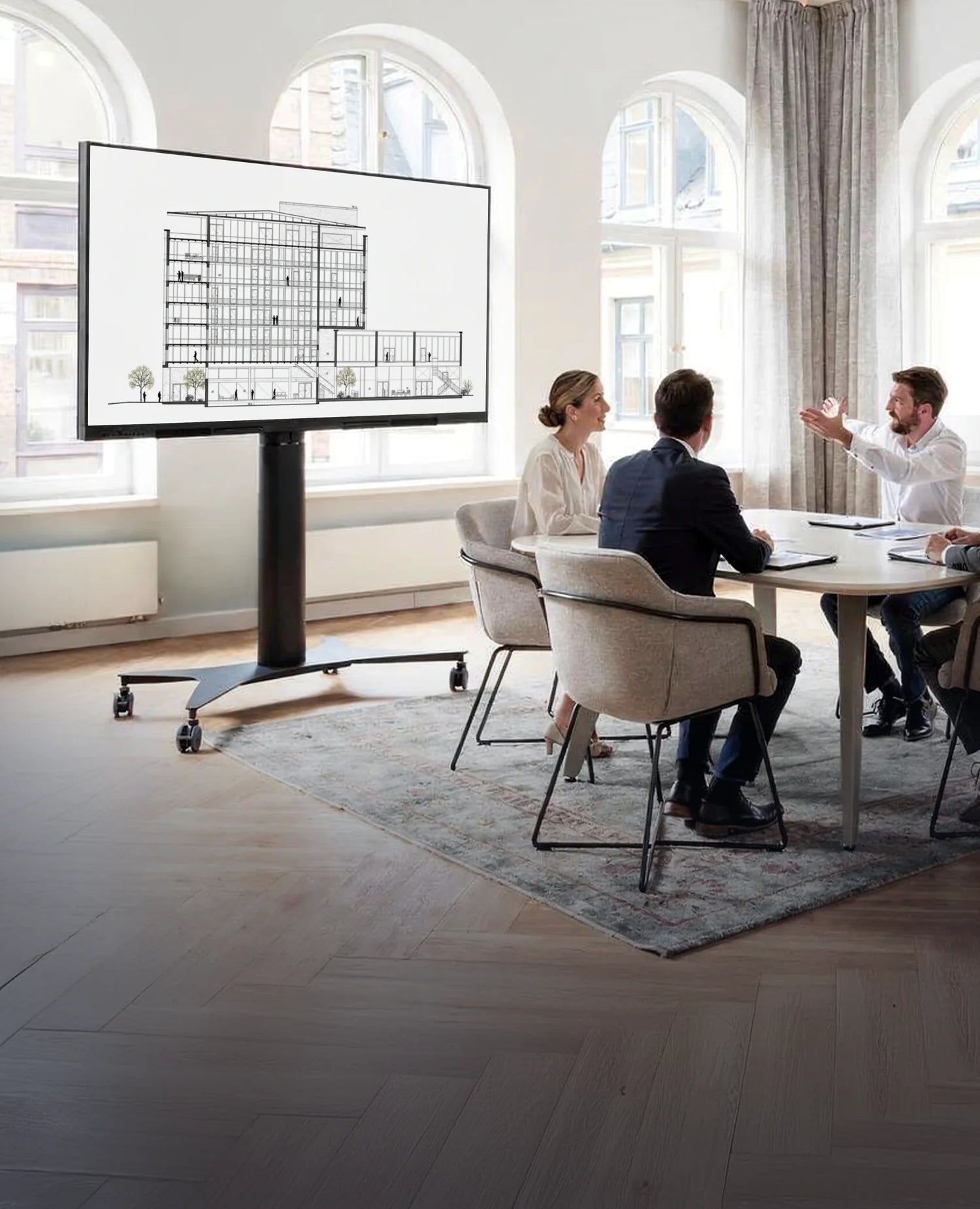Four people are seated at a table in a bright, modern room, holding a meeting. On the left stands a celexon Expert electrically height-adjustable display trolley Adjust 42100M, showing an architectural drawing.