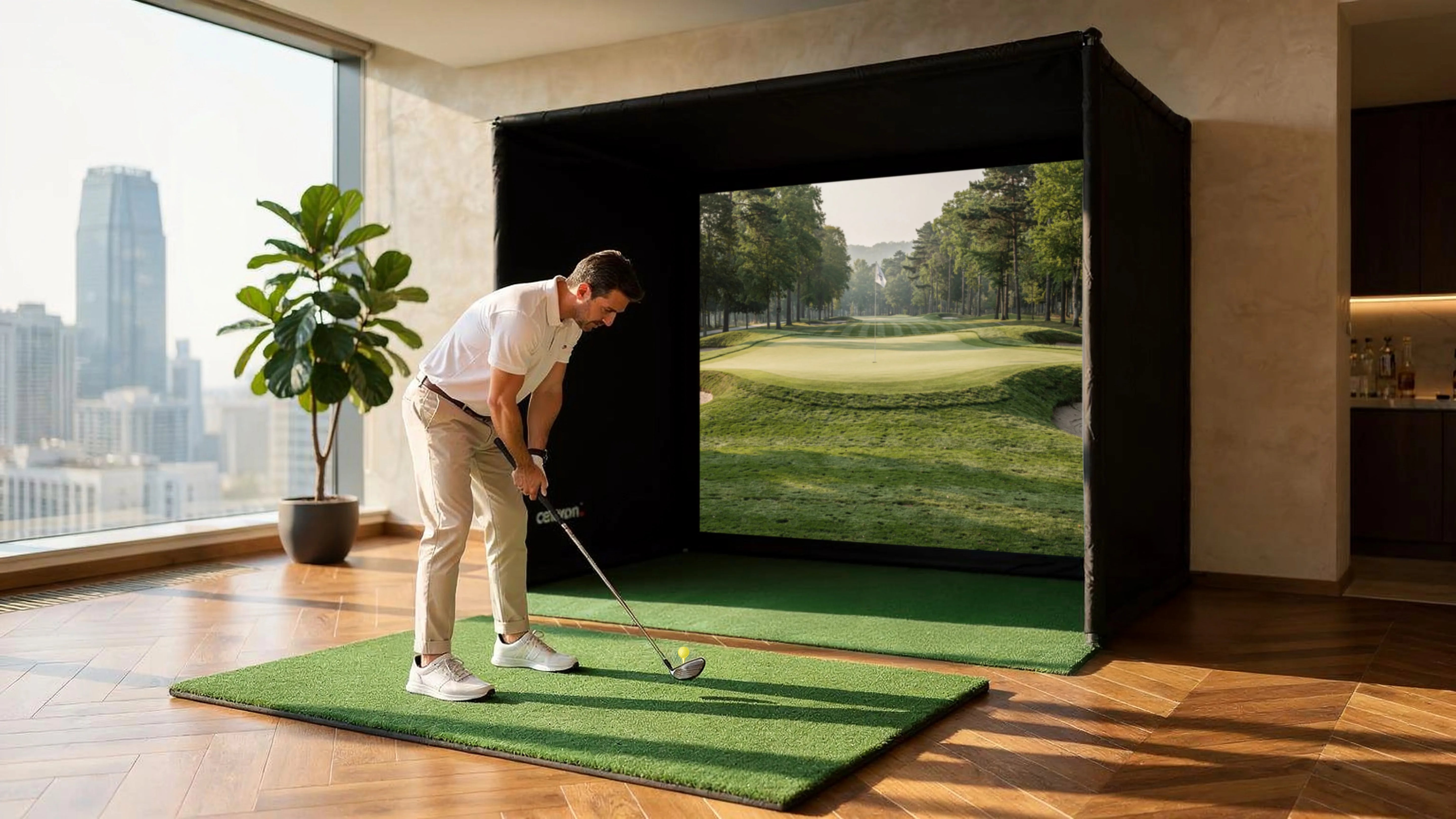 The image shows a man practicing on an indoor golf simulator in a modern apartment. He stands on a small artificial turf mat, preparing to take a swing, while in front of him a large projection screen inside a black simulation enclosure displays a golf course with a fairway and green.