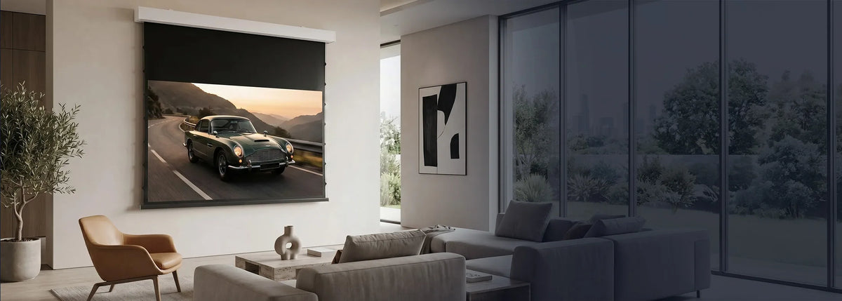 A modern living room featuring a celexon DARKVISION ALR Premium Plus Screen Electric Tension, displaying a classic sports car on a winding mountain road. Light-colored seating, a leather armchair, and a large indoor plant complement the elegant, minimalist interior.