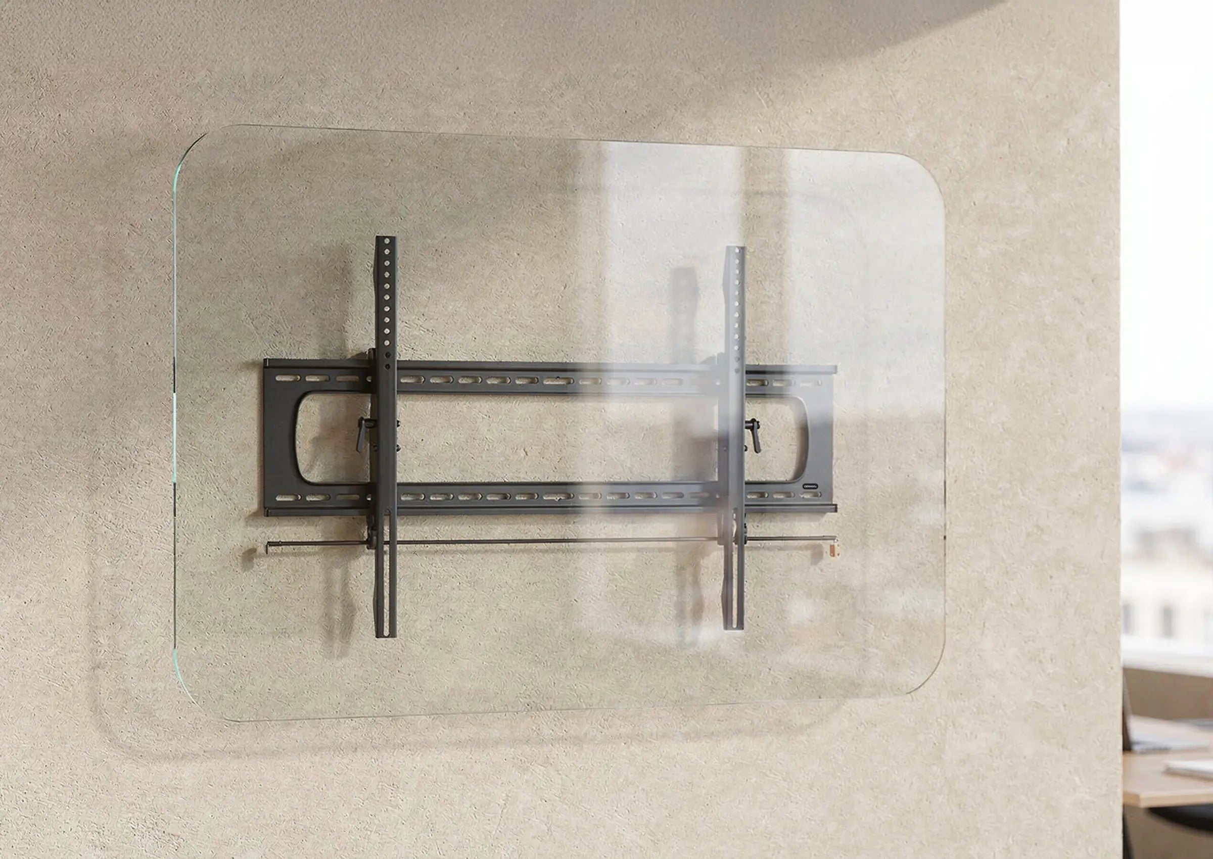 The image shows the celexon TV/display wall mount Adjust/Fixed attached to a transparent glass panel. The bracket consists of a sturdy metal construction with horizontal and vertical rails for mounting a screen. The scene is set in a modern office or workspace with a neutral wall, large windows, and a desk in the background.