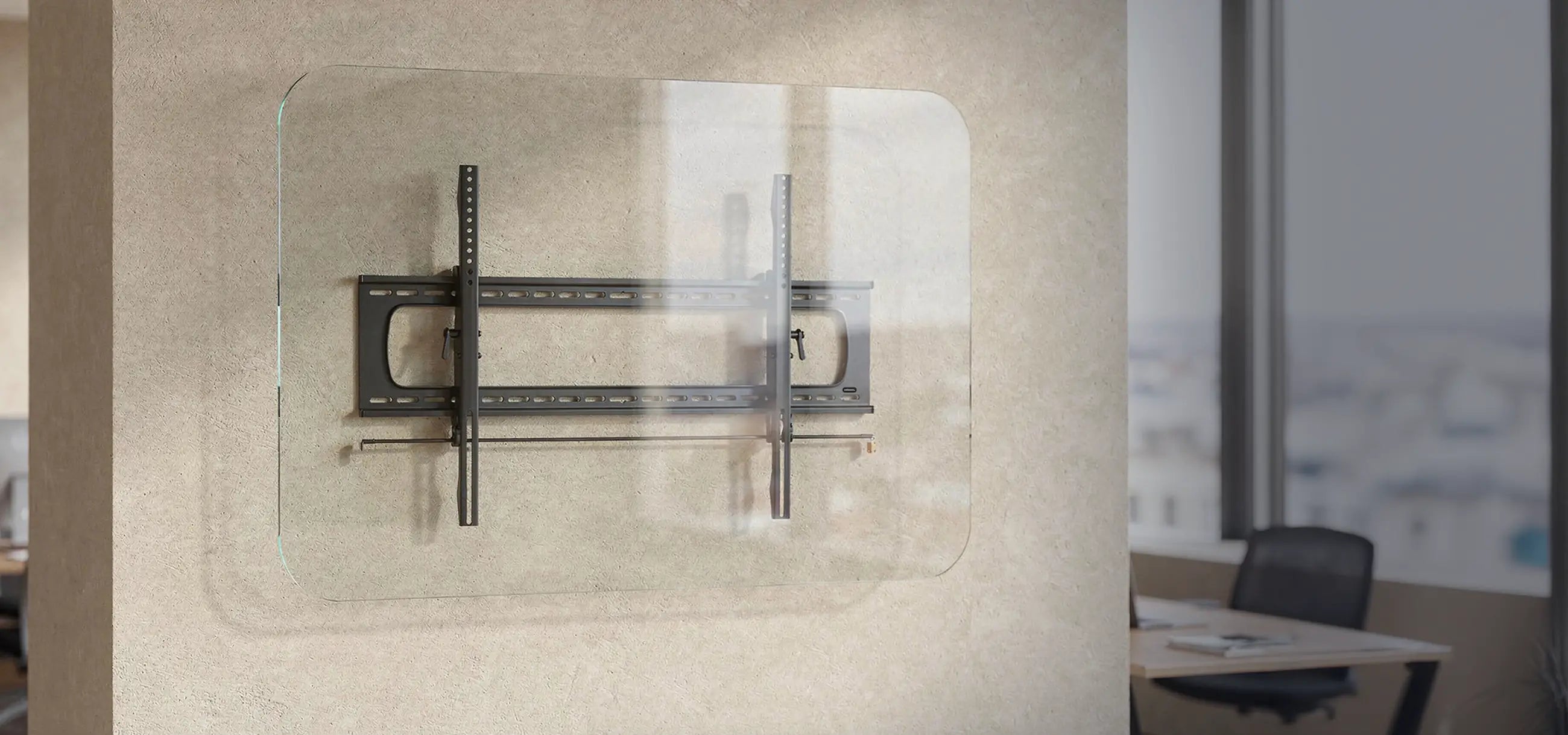 The image shows the celexon TV/display wall mount Adjust/Fixed attached to a transparent glass panel. The bracket consists of a sturdy metal construction with horizontal and vertical rails for mounting a screen. The scene is set in a modern office or workspace with a neutral wall, large windows, and a desk in the background.