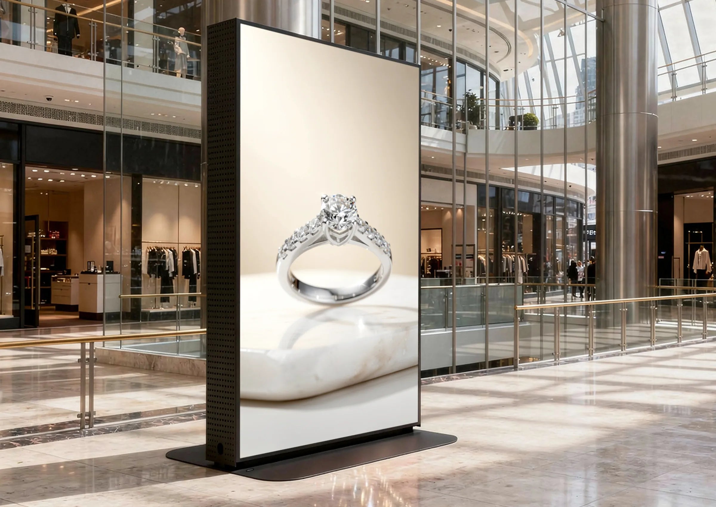 The image shows a large, freestanding digital advertising display in a modern shopping mall with high glass architecture and open galleries. On the vertical screen, an elegant advertisement of a diamond ring is presented. Boutiques and shop windows surround the display.