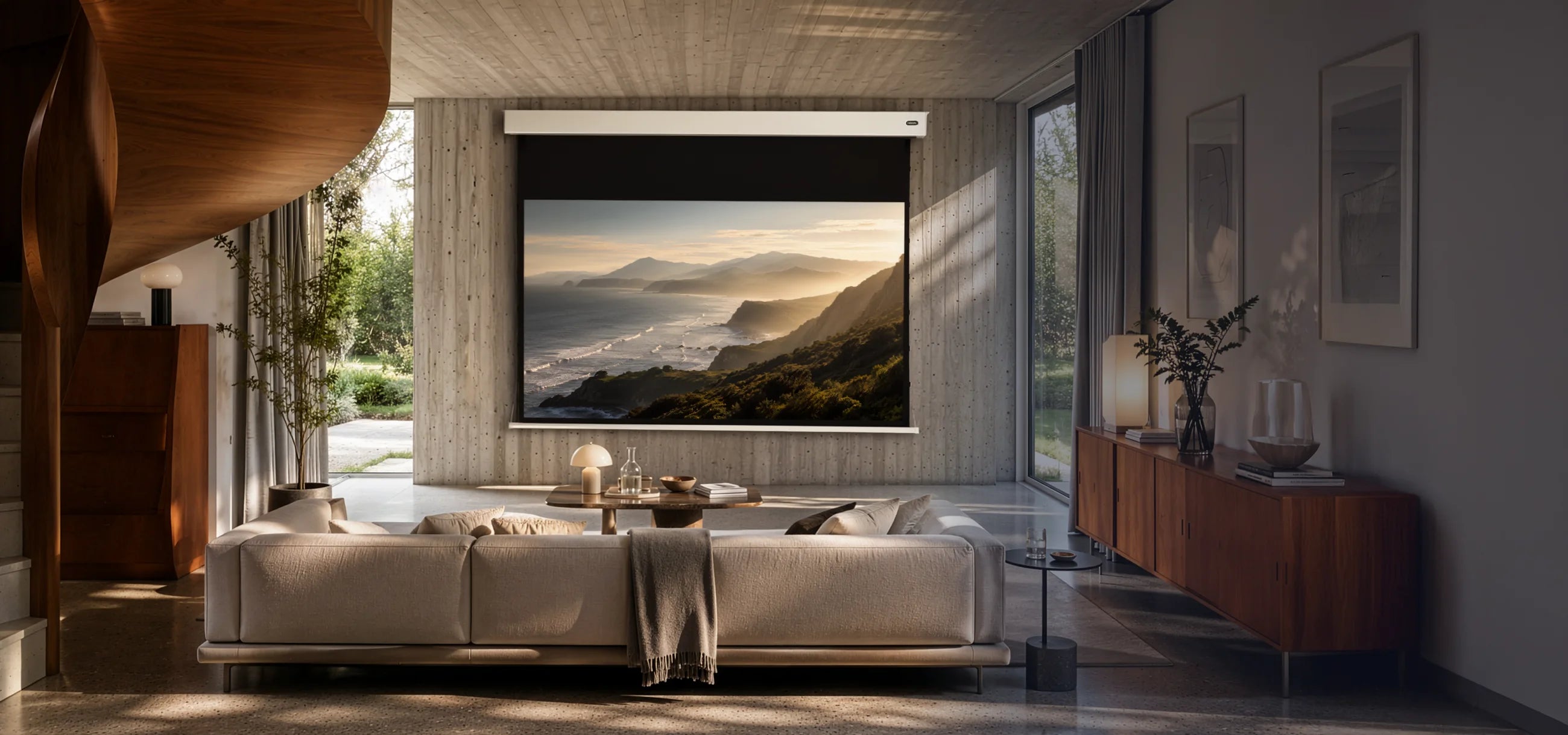 A modern lifestyle presentation of a celexon Pearlstone projection screen in a premium living space. Front view with the screen fully extended, displaying a warm coastal projection. Exposed concrete, natural wood accents, floor-to-ceiling windows, and soft natural daylight create an elegant, high-end atmosphere with a clear focus on the product.