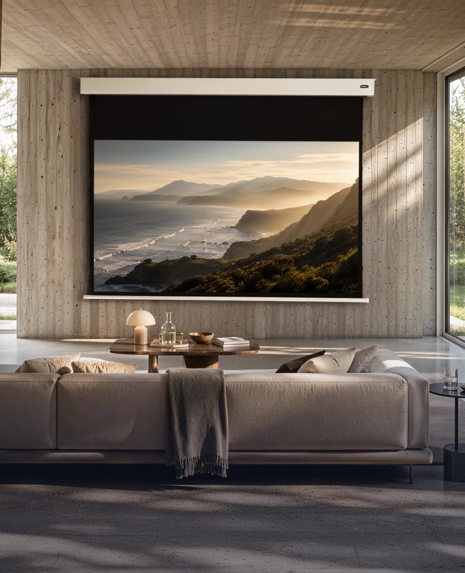 A modern lifestyle presentation of a celexon Pearlstone projection screen in a premium living space. Front view with the screen fully extended, displaying a warm coastal projection. Exposed concrete, natural wood accents, floor-to-ceiling windows, and soft natural daylight create an elegant, high-end atmosphere with a clear focus on the product.