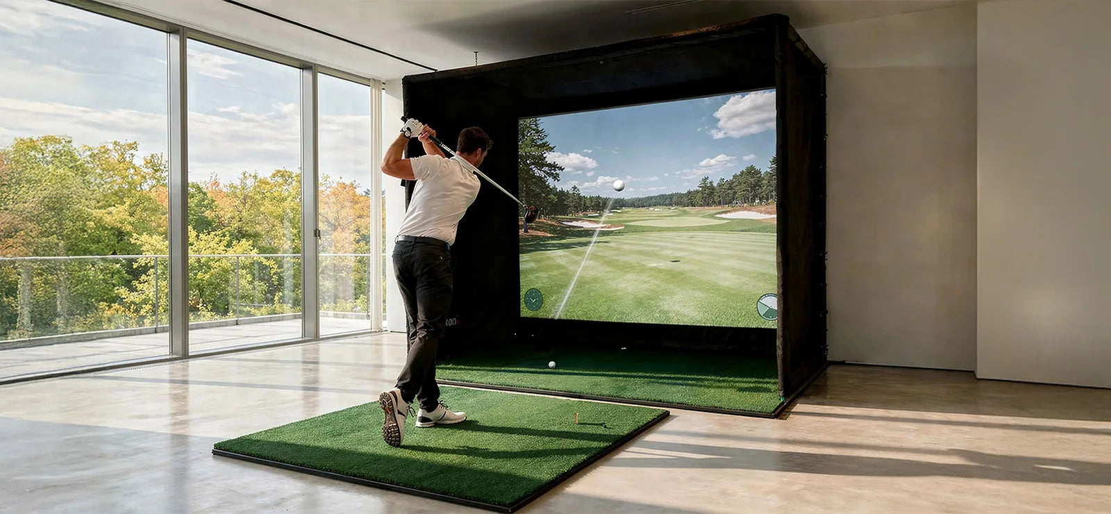 The image shows a man in a modern room taking a swing on an indoor golf simulator. He stands on an artificial turf mat and has just swung his golf club, while the large projection screen in front of him displays a virtual golf course with the trajectory of the ball.
