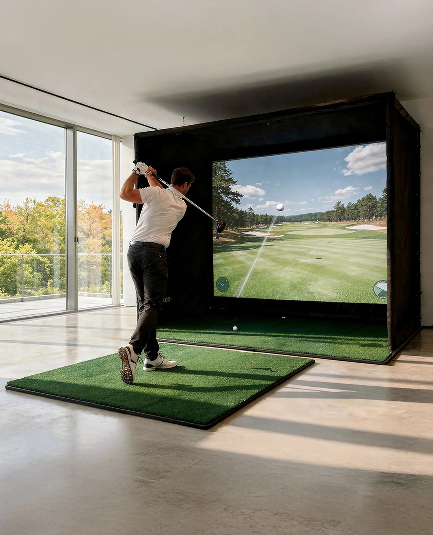 The image shows a man in a modern room taking a swing on an indoor golf simulator. He stands on an artificial turf mat and has just swung his golf club, while the large projection screen in front of him displays a virtual golf course with the trajectory of the ball.