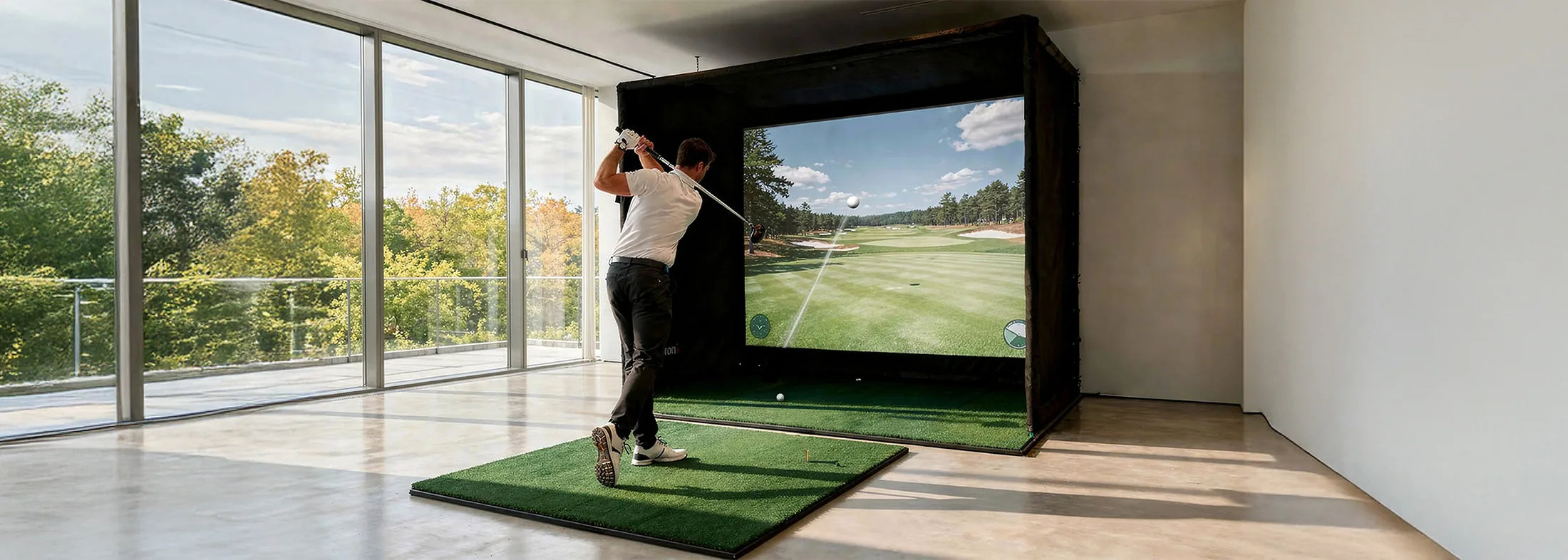 The image shows a man in a modern room taking a swing on an indoor golf simulator. He stands on an artificial turf mat and has just swung his golf club, while the large projection screen in front of him displays a virtual golf course with the trajectory of the ball.