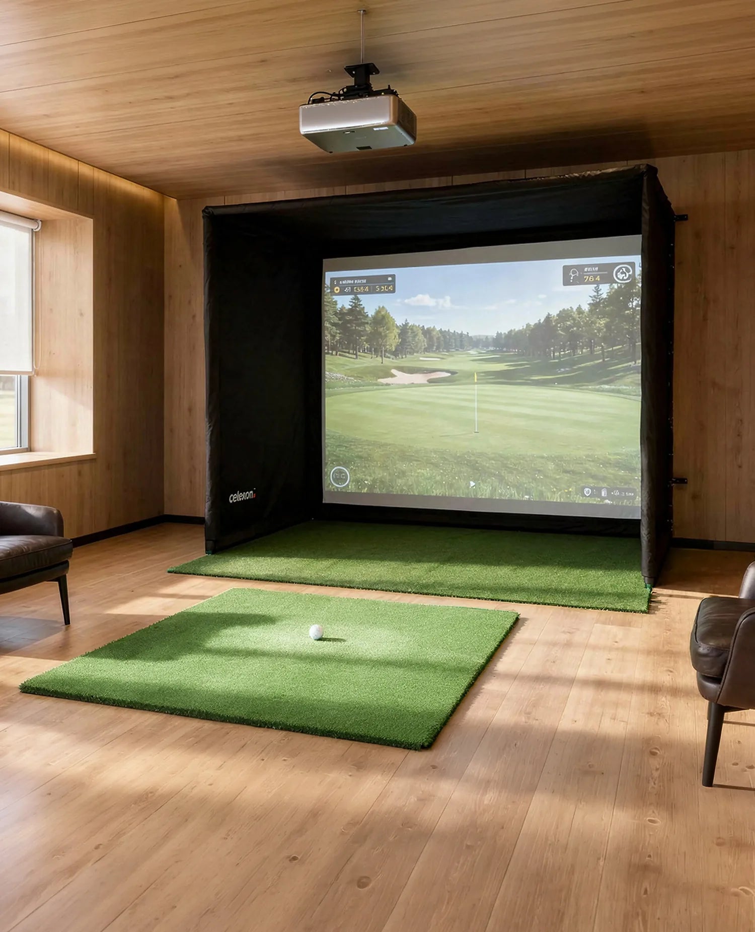 The image shows a stylish indoor golf simulator in a modernly furnished room with warm wood paneling. In front of a large projection screen inside a black simulation enclosure lies an artificial turf mat with a golf ball ready, while a projector mounted on the ceiling projects the virtual golf game onto the screen. Several golf clubs stand next to the setup, and two comfortable leather armchairs as well as large windows with a view outside complete the scene.