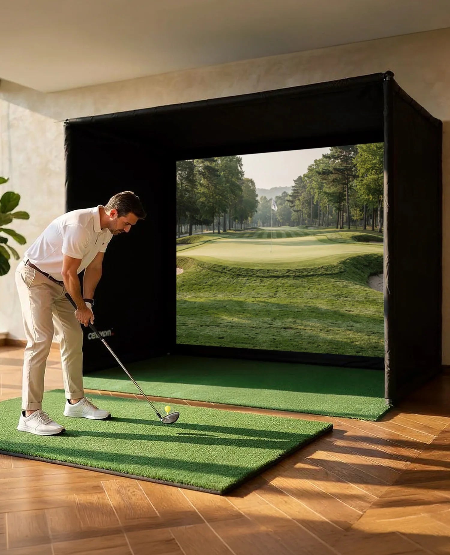 The image shows a man practicing on an indoor golf simulator in a modern apartment. He stands on a small artificial turf mat, preparing to take a swing, while in front of him a large projection screen inside a black simulation enclosure displays a golf course with a fairway and green.