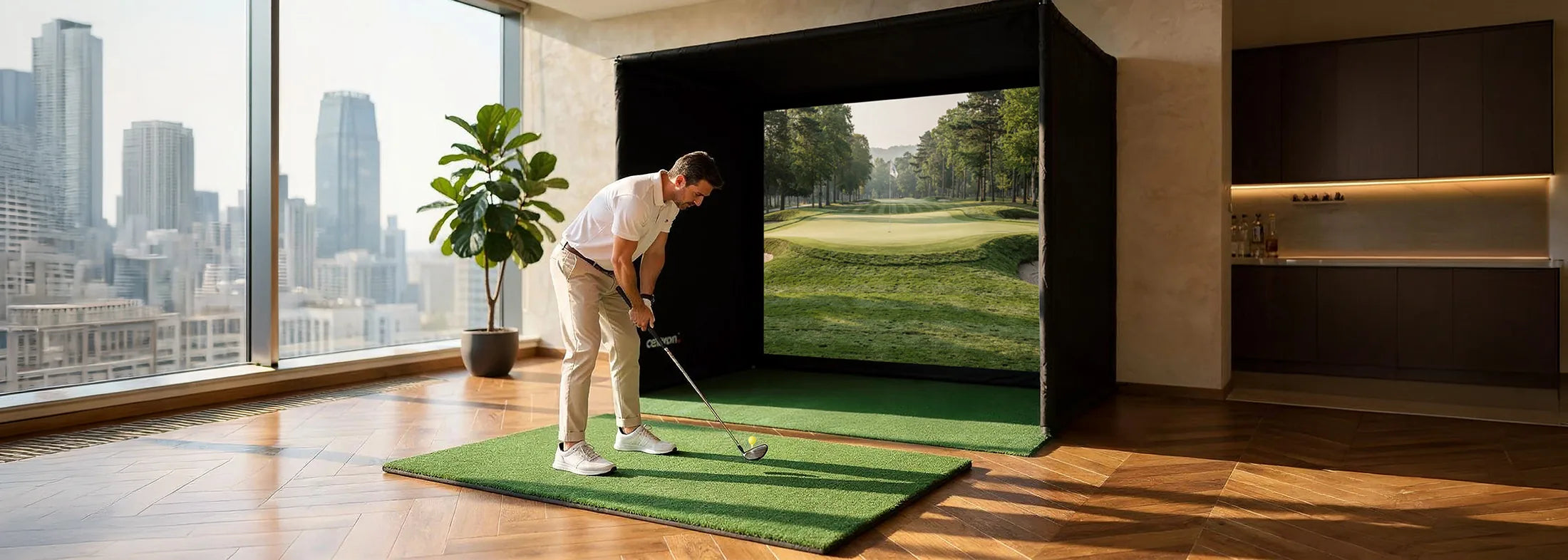 The image shows a man practicing on an indoor golf simulator in a modern apartment. He stands on a small artificial turf mat, preparing to take a swing, while in front of him a large projection screen inside a black simulation enclosure displays a golf course with a fairway and green.